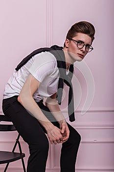 Modern fashion model of a young man in glasses in a stylish white t-shirt in fashionable jeans with a trendy hairstyle sits on a