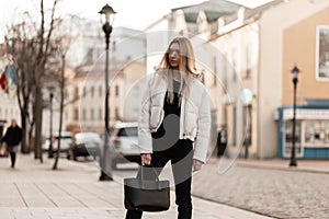 Modern fashion model of a young woman in a white fashionable jacket in stylish jeans with a leather trendy bag stands on the