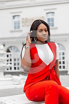 Modern fashionable young black woman in beautiful stylish red suit is resting on wooden bench in city. Glamorous trendy african