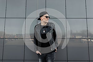 Modern fashionable young hipster man in stylish black clothes in a black baseball cap in sunglasses relaxes standing