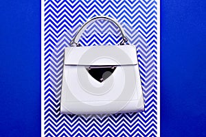 Modern female top-handle bag from white leather with black trim, metal detail, and feet isolated on a blue-white background with a