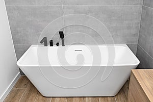 A modern, free standing wall mounted bathtub with a black matt tap, standing in a bathroom lined with ceramic tiles.