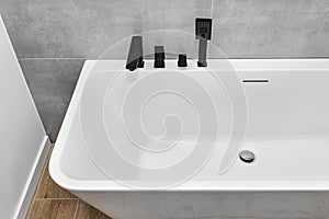 A modern, free standing wall mounted bathtub with a black matt tap, standing in a bathroom lined with ceramic tiles.