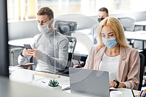 Modern gadgets for working with clients in office. Millenial man in mask typing on smartphone, lady works in laptop at