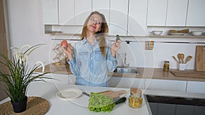 Modern girl chooses from which vegetable to prepare salad and smiles into camera, standing in middle of kitchen at