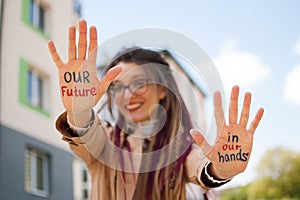 Modern girl with long dreadlocks and eyeglasses in nude color trench is showing hands with written slogan `Our future in