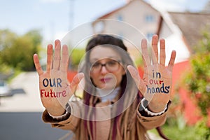 Modern girl with long dreadlocks and eyeglasses in nude color trench is showing hands with written slogan `Our future in