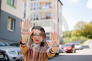 Modern girl with long dreadlocks and eyeglasses in nude color trench is showing hands with written slogan `Our future in