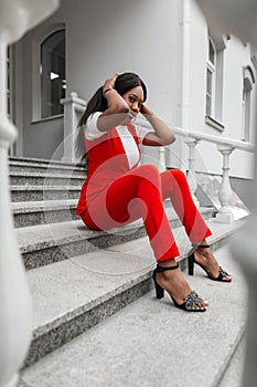 Modern glamorous black young woman with sexy lips in fashionable red-white clothes in fashion shoes rests on vintage stone
