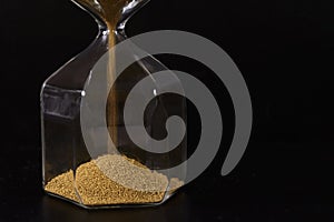 Modern glass hourglass with yellow sand on black background, close-up, copy space