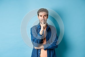 Modern guy shushing at camera, press finger to lips and telling a secret, making taboo sign, standing on blue background