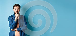 Modern guy shushing at camera, press finger to lips and telling a secret, making taboo sign, standing on blue background