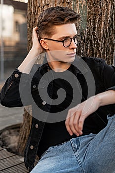 Modern handsome young man in stylish jeans clothes straightens trendy hairstyle outdoors. Urban guy in vintage casual denim wear