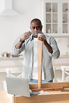 Modern handyman and online instructions. Black man screwing detail with manual screwdriver to table leg in kitchen