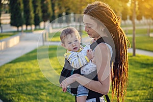 Modern happy mother with baby son in ergo backpack walking in Sunny summer day. Concept of the joy of motherhood