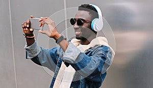 Modern happy smiling young african man taking selfie picture by smartphone while listening to music in wireless headphones on gray