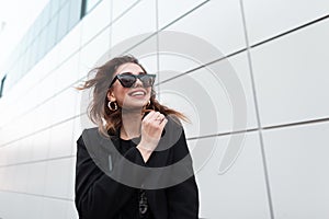 Modern happy young hipster woman in a black stylish coat in dark trendy sunglasses goes and smiles around the city