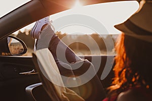 Modern hipster girl resting in a car and reading a map. Woman with feet on car door. Feet outside the window at sunset