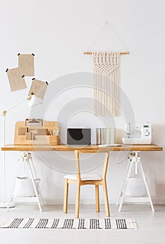 Modern home offfice interior with a desk, laptop, sewing machine, chair and macrame on a wall. Empty screen, place