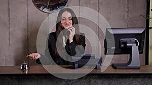 Modern hotel reception desk with bell. Happy female receptionist worker standing at hotel counter and talking to phone.