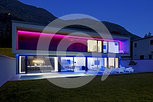 Modern house with swimming pool and garden in night scene illuminated by colored LED lights. Behind the house is the hill with the