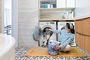 Modern housewife putting dirty linen textile and clothes to washing machine at domestic laundry