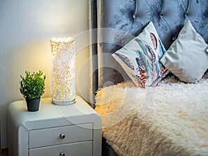 Modern interior of bedroom. Burning lamp on white nightstand.