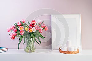 Modern interior design with tulip flowers bouquet in vase, white picture frame mockup, candles on white console on pink