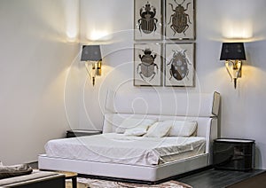 Modern interior, white bedroom with a double bed, paintings and lamps on the walls. Beautiful design of the bedroom