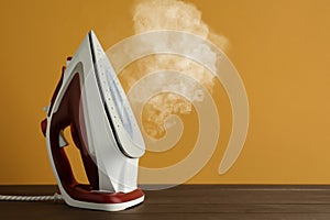 Modern iron with steam on wooden table against orange background, space for text