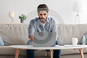 Modern job. Concentrated arab man working on laptop at home office, sitting on sofa and using computer
