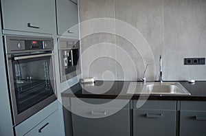 A modern kitchen corner interior with a gray tiled wall, kitchen sink, black electrical outlets and counter, gray cabinets, and