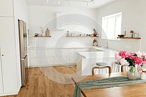 Modern kitchen interior. Beautiful peonies in vase on wooden table and stylish white kitchen with appliances in new scandinavian