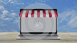 Modern laptop computer with online shopping store graphic and open sign on wooden table over blue sky with white clouds, Business