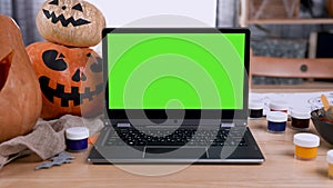 Modern laptop with green screen mockup with chroma key stands on the table in the room. Next to the laptop are pumpkins