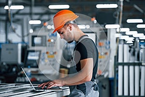 Modern laptop. Typing on keyboard. Industrial worker indoors in factory. Young technician with orange hard hat