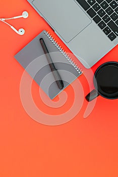 Modern laptop, white headphones, gray notebook with a pen and a cup of coffee on the red background