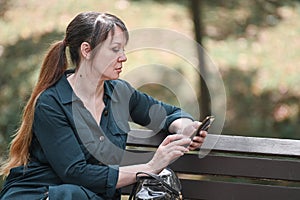 Modern lifestyle concept. Serious mature woman sitting on a park bench using smartphone, messaging via online application