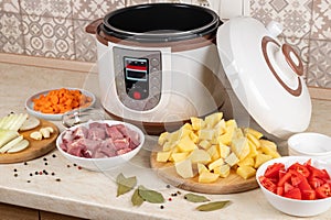 Modern, light multi cooker and products on table in kitchen. Cooking stewed potatoes with meat in multicooker