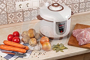 Modern, light multi cooker and products on table in kitchen. Cooking stewed potatoes with meat in multicooker