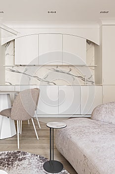 Modern living room interior in pastel colours with round dining table, beige chairs, creamy white kitchen furniture