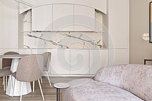 Modern living room interior in pastel colours with round dining table, beige chairs, creamy white kitchen furniture.