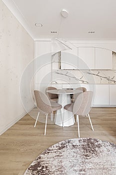 Modern living room interior in pastel colours with round dining table, beige chairs, creamy white kitchen furniture.
