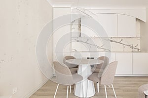 Modern living room interior in pastel colours with round dining table, beige chairs, creamy white kitchen furniture.