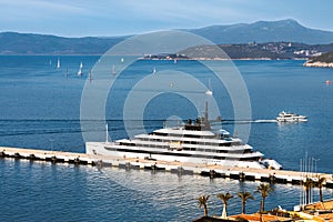 A modern luxury yacht in a Mediterranean port in the background lots of sailboats and mountains