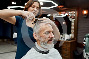 Modern male hairstyle. Handsome bearded mature man sitting in armchair in the barbershop while young barber girl doing