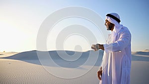 Modern male sheikh uses smart watch and stands in middle of bottomless desert under scorching sun in morning.