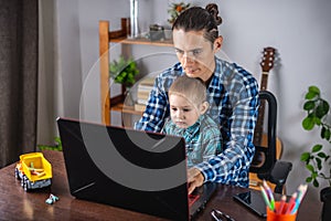 Modern man is working on a laptop, and his little son is sitting on his lap. Concept of family and remote work from home