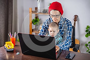 Modern man is working on a laptop, and his little son is sitting on his lap. Concept of family and remote work from home