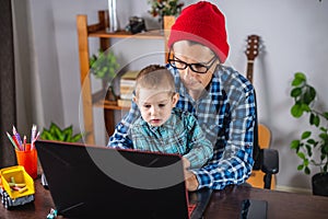 Modern man is working on a laptop, and his little son is sitting on his lap. Concept of family and remote work from home